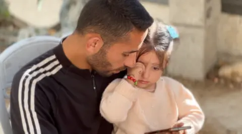 Julia rubs one of her eyes with a hand as she leans against her father, who holds her on his lap. Mohammed is a young man with dark hair and a trimmed beard. They are sitting down in a plastic chair, outside.