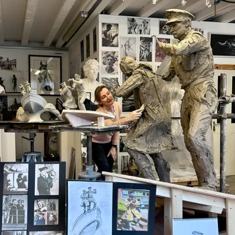 Amy Goodman is pictured in her workshop, using a tool on a statue in progress. Near her are figurines of two men on the prow of a boat, showing how part of the Coastal Forces memorial will look. Photographs of Royal Navy Wrens are positioned in front of her.