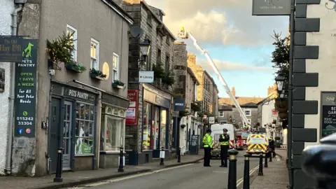 Emergency services on Market Street in Kirkby Lonsdale. It is a narrow road with older brick buildings on either side. Two police officers stand in the road. Behind them are emergency vehicles and, in the distance, a firefighter is standing in an extended cherry-picker.