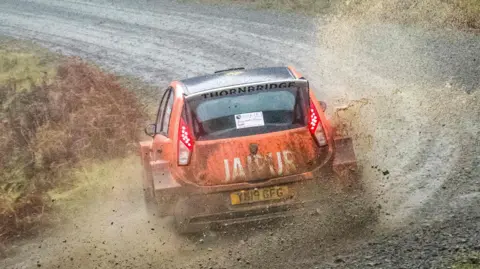 Rally Media UK An orange rally race car travels at speed on a gravel track. The car has sponsorship stickers plastered across the side of the car, one reading 'JAIPUR'. The vehicle is travelling in a forest. 