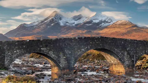 Steve Gunter A bridge is in the foreground, with snow capped hills and mountains in the background, and sunlight across the scene