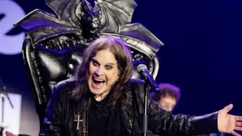 Ozzy is sat on his throne on stage at his farewell gig last year. The chair is black and he is wearing black with a cross necklace on his chest. His hair is He is waving his left hand. dark and shoulder length.