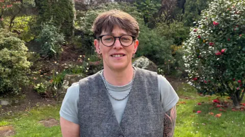 A woman with glasses smiling at the camera, wearing a grey vest jacket