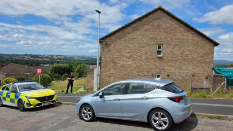 BBC A police car and officer stand outside a row of brick terraced houses cordoned off with tape in a residential street, as officers attend a property. A light blue car is also visible parked in front of the police car. 