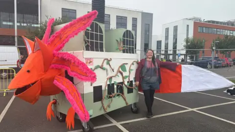 A woman standing next to a carnival float, which has a red creature with the tail of a fox and the head of an axolotl protruding from a square cardboard representation of an incinerator.