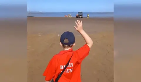 A boy standing on the beach wearing a red shirt with his arm up