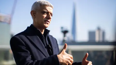 PA Media Sir Sadiq stood on a rooftop with high rise buildings blurred in the background. He is wearing a dark navy coat and gesticulating/ talking to someone to the right of the camera.