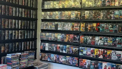 Rachael McMenemy/BBC DVDs are piled high on top of shelves and on display in large black shelves on the wall.