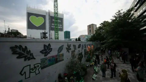 Reuters A large group of people gather at the memorial wall where flowers arer laid. It is in front of the remnants of the Grenfell tower block, which are wrapped in white plastic material and a sign reading 'Grenfell Forever in our hearts'.