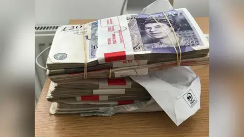 CPS A stack of £20 notes with paper and elastic bands around them, on a wooden desk. 