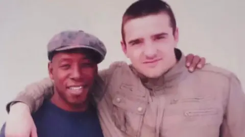 Family photo Footballer Ian Wright is wearing a cap and a blue top. He has his arm around a taller man wearing a beige jacket who has short brown hair.