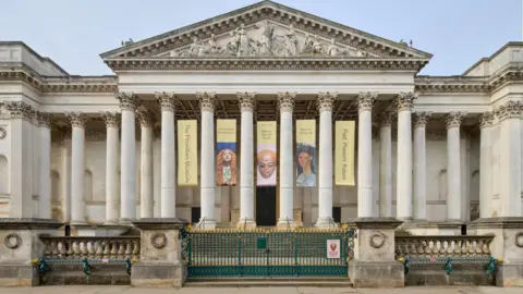 Front view of the University of Cambridge's Fitwilliam Museum. It is an ornate building with columns and scuptures on the top. It has green and gold gates at the entrance.