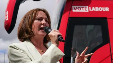 Lola McEvoy speaking into a microphone at an election campaign event in 2024.  Behind her is a red bus with "Vote Labour" branding on the side.