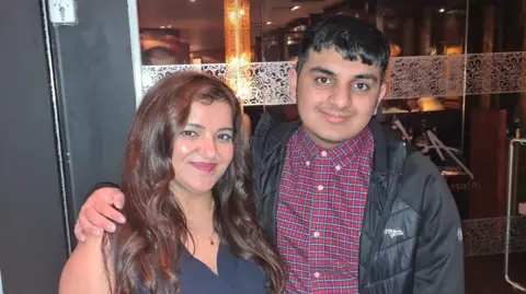 Family Pooja and her son Ronan are stood together outside a restaurant. Ronan has a black jacket on and checked shirt and has his arm around his mother who has a navy sleeveless top on.