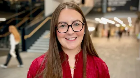 Rachel Huddleston - a young woman with long brown hair smiles at the camera. She is standing in the middle of the university campus, but the background is blurred. She is wearing black-framed square glasses, a red shirt and a gold necklace.