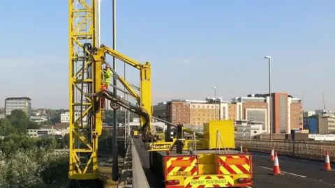 Gateshead Council A highway maintenance vehicle parked on the Redheugh Bridge between Newcastle and Gateshead. The bright yellow vehicle holds a large piece of equipment which is stretched over the side of the bridge. The equipment holds a large ladder which is standing vertical off the side of the bridge. A man in a hi-vis jacket is climbing on to the ladder.