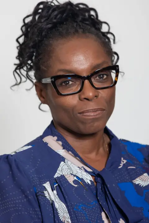 Grainge Photography Dr Lade Smith, wearing black-framed glasses and a blue top, looks directly to camera.