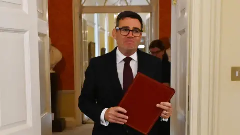 Andy Burnham clutches a red folder as he walks through an open doorway in 10 Downing Street. He is wearing a dark suit and tie