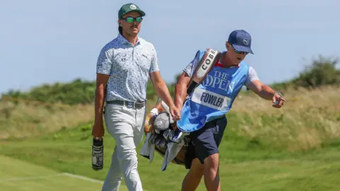 Pacemaker Rickie Fowler and his caddie walking along the course. He is wearing green sunglasses and a green cap. His caddie is wearing a bright blue bib with the name Fowler across the front.