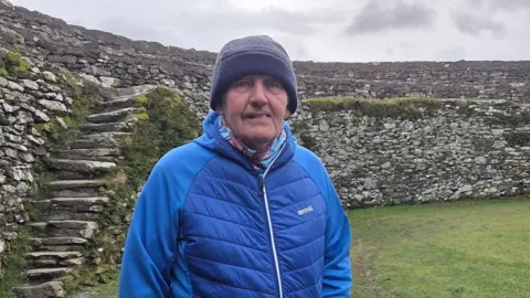 A man in a blue coat and hat stands in the middle of An Grianán of Aileach, a stone ringfort in County Donegal, Ireland.