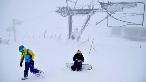 Two snowboarders in whiteout conditions at Cairngorm Mountain. There is ski lift infrastructure in the background.