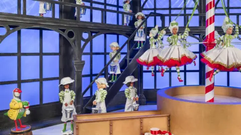 Andrew Meredith Foggy the partridge is in the corner wearing yellow, as well as eight maids wearing white and holding milk pails. There are nine women dancing around a red and white striped pole.