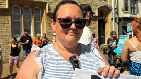 Kelsey Day wearing sunglasses and a white and grey striped top, standing among a crowd of protesters