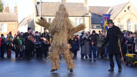 A man dressed as a straw bear is in the middle of the image, jumping. People are stood in a semi-circle behind and looking. 