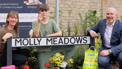 Vox Online Image shows Fiona, Will and Chris Parker with the new Molly Meadows sign, with some cat treats at the base of the sign. 