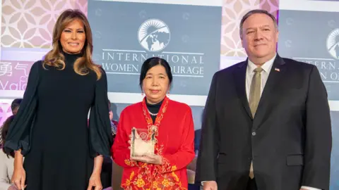 US Department of State Susanna Liew is wearing an elaborate red and yellow embroidered jacket and holds up a glass trophy, smiling slightly, as she stands on stage between Melania Trump who is wearing a black dress and Mike Pompeo who is wearing a black suit and gold tie.
