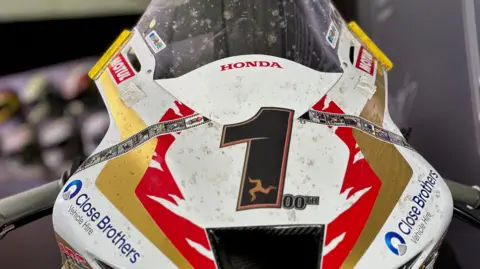 The front of the machine, which is white red and gold, with a large number one on the front in black that has the three legs of Man in gold and a smaller 00th next to it.
