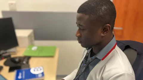 Benjamin Andoh is sitting at a desk, wearing a white coat and an NHS lanyard. He is looking away from the camera, probably at a computer which has been cut out of the photo.