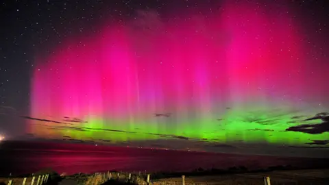 BBC Weather Watchers/Saf37y A picture of the night sky across a big empty expanse. The sky is lit up bright green and pink in waves, showing the aurora, in front of a sparkling night sky filled with stars