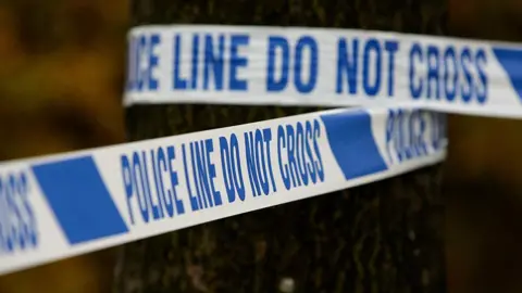 Generic image of police tape wrapped around a tree, it says 'police line do not cross' in blue writing on a white background