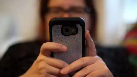 The image shows a mobile phone in the foreground hiding the face of the user behind it.