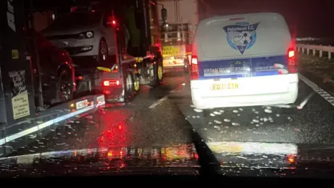 Owen Clanachan A photograph taken from inside a car. Traffic on the road outside is at a standstill. There is a small white van in front and a car transporter to the left. It is dark and wet. 