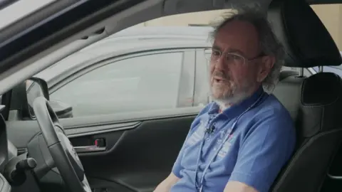 Turner photographed sat at the wheel of his car, looking out of the passenger window. He is wearing a blue polo shirt with 'Shrewsbury and Telford Hospital Trust' and 'Volunteer' embroidered on it in white lettering. He has short, grey, curly hair, a beard, and is wearing thin-framed glasses. The interior of his car is black. 