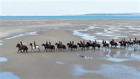 A drone shot of dozens of cavalry horses in a line formation walking along the sand with the sea in the distance on Hayling Island beach