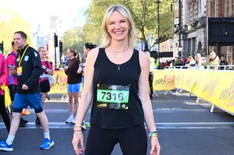 PA Media Jo Whiley wearing a black running top and leggings standing among other runners surrounded by yellow hoardings