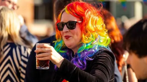 Sorted PR A smiling woman wearing sunglasses, a rainbow coloured wig and dark jacket holds aloft a drink.
