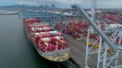 Getty Images A wide shot of a container ship docked at a container port with a grey crane in the foreground