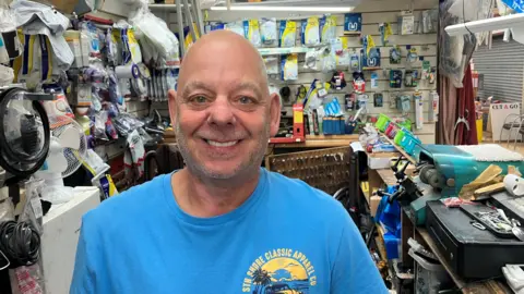 Photograph of Anthony Ainscough, who runs Vacuum Repairs at Wigan Indoor Market. The image shows him inside his store, with his stock in the background.