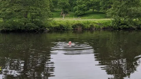 Jo Broughton A woman swimming in a river by a tow path with greenery surrounding the bank