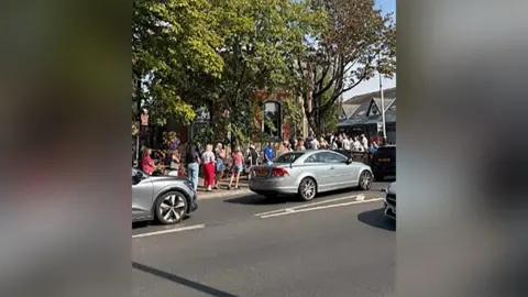 Handout An image of the back of a long queue of people waiting to buy eclipse glasses from Specsavers in Formby. It is taken from across the road, with cars on the road in the foreground.
