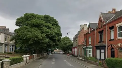 Elwick Road has two-storey red-brick terraced housing on the right and beige brick two-storey terraced housing on the left. There are a couple of mature trees in the front gardens of houses on the left, and hedges in some of the smaller front town gardens on the right.