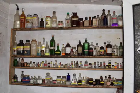 Bedfordshire Police Shelves of varying heights within a wooden surround, mounted a white brick wall. There are dozens of bottles on three shelves within the surround, and more on top of the unit.