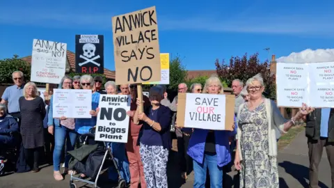 A group of campaigners standing along a residential street holding up signs. They read 'save our village', 'Anwick village says no' and 'Anwick says no to biogas plant'.