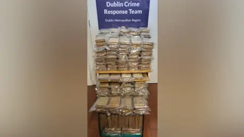 Gardaí Handout A still image showing numerous brown mid-size parcels sealed in plastic bags. The packages are stacked on top of one another, with some stored in a green crate on the ground. They are placed in front of a large blue poster which reads 'Dublin Crime Response Team' in white font.