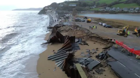 The A379 Slapton Line between Torcross and Slapton has washed away. The image shows a gaping hole in the road, bent metal sea defences, red barrier and some heavy plant machinery.