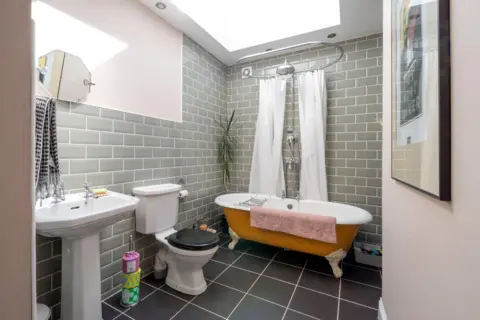 IWC Media A bright, modern bathroom features pale green subway tiles, a skylight, and a freestanding bathtub with a curved shower rail and white curtain.
A white pedestal sink, toilet, and simple accessories sit against dark floor tiles, while soft daylight fills the compact space.